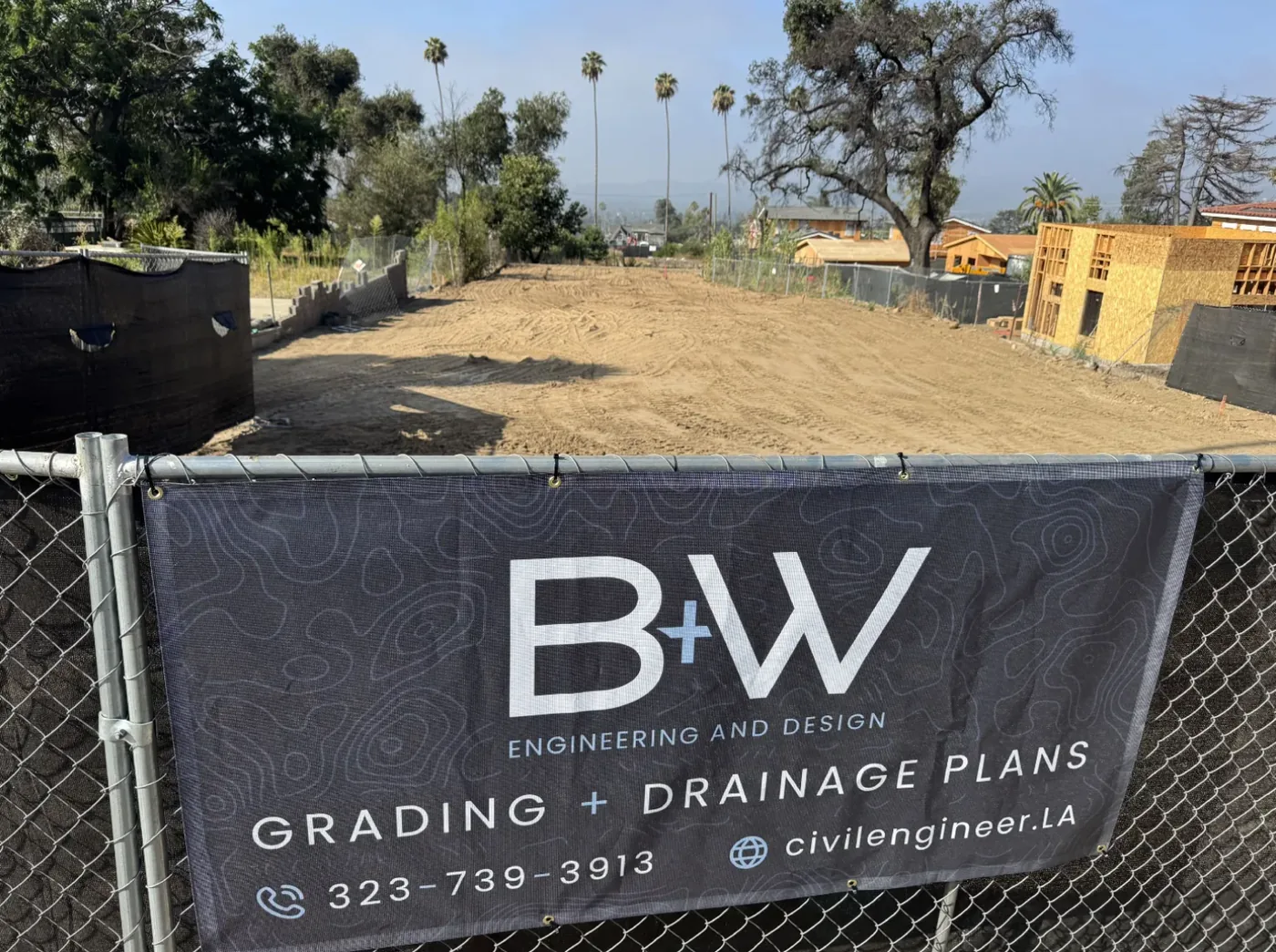 B+W Engineering mesh banner zip-tied to a chain link fence at a graded residential rebuild lot in Altadena, with framed houses under construction behind it